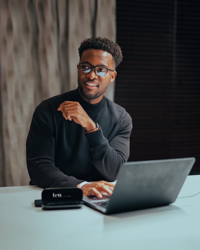 Cheerful man with glasses working on laptop at a table indoors, emanating a professional and modern workspace vibe.