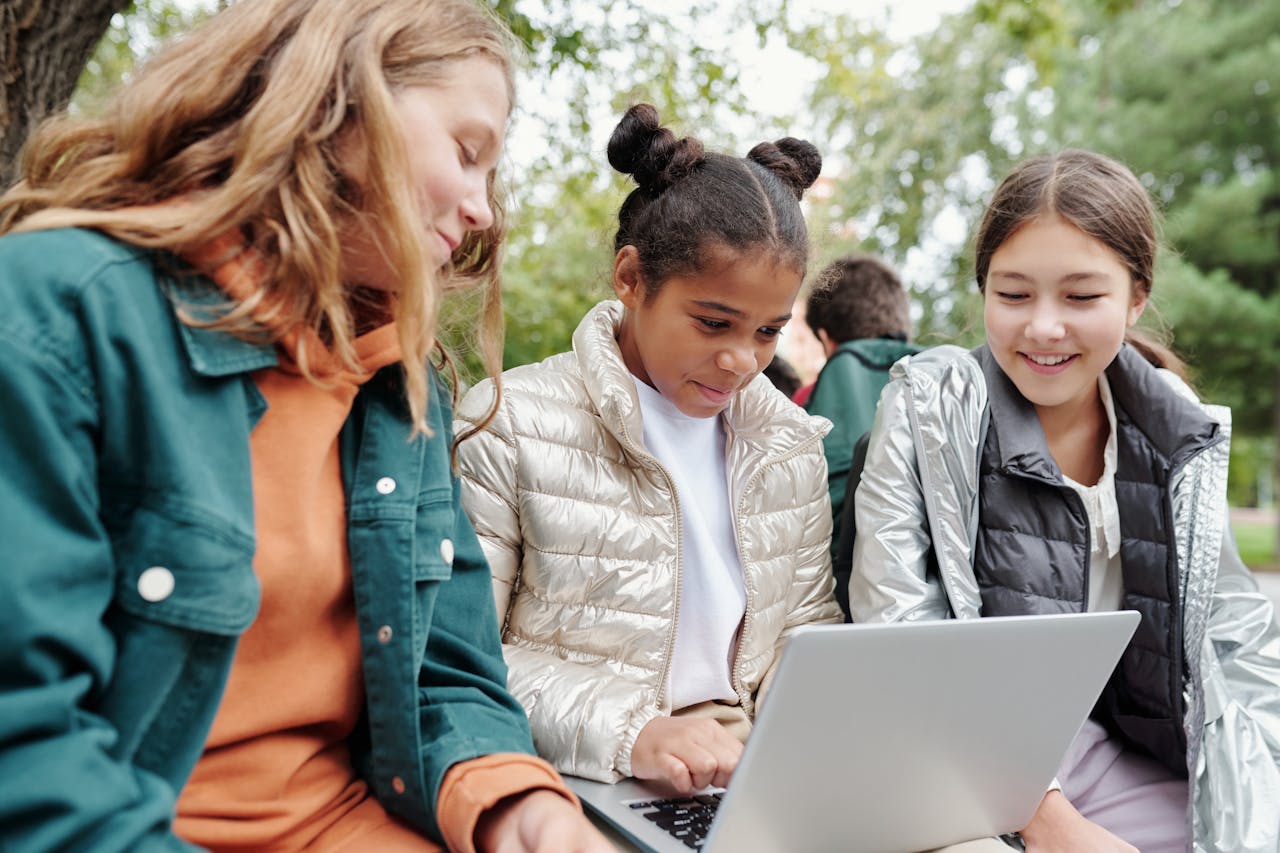 Home Three girls sitting together outdoors, smiling and learning on a laptop in a park setting.